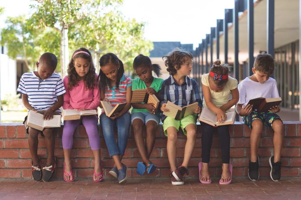 Front,View,Of,Diverse,Students,Reading,Book,While,Sitting,On brick